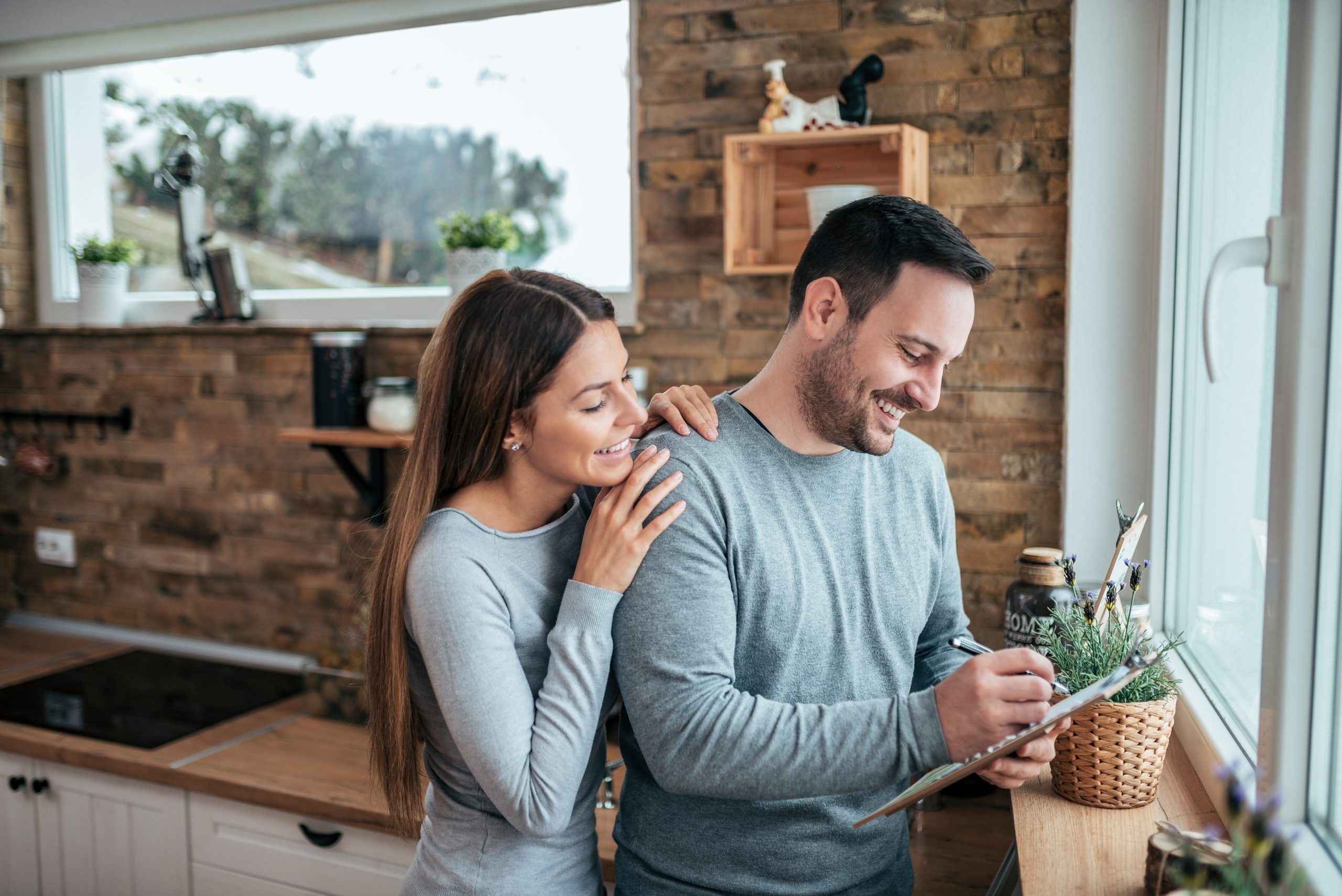 Cute Couple Standing In The Kitchen Near The Window And Man Writing On Paper.